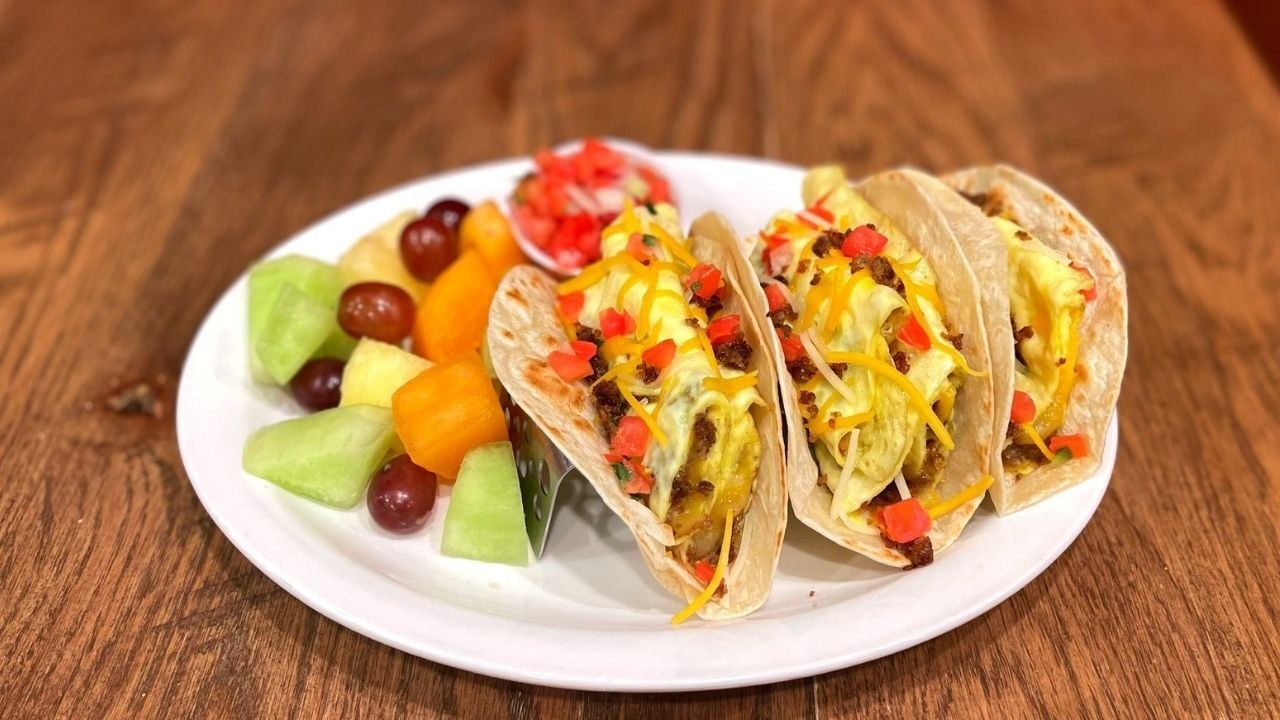 A white plate with 3 breakfast tacos and a side of fruit.
